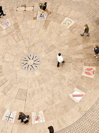 Several people walk across a square in which coats of arms are embedded in the ground.